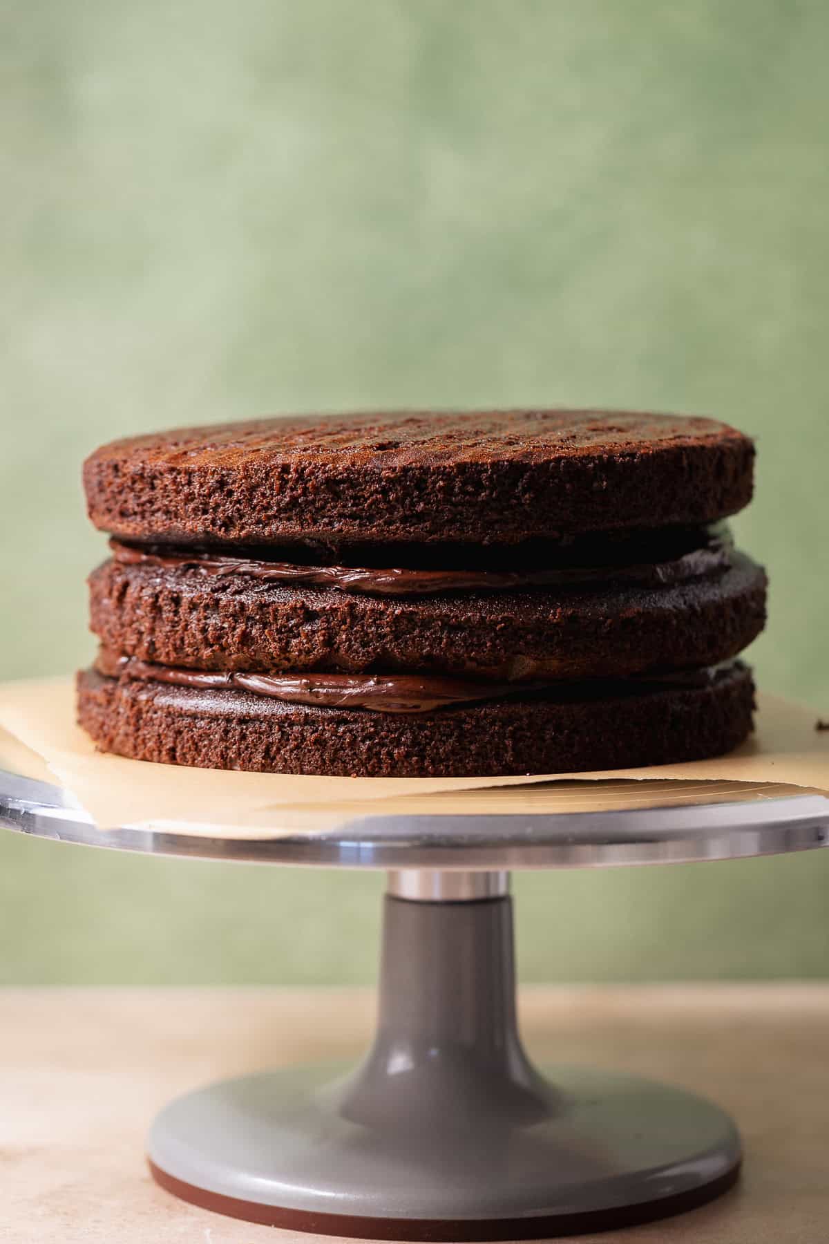 Three chocolate cake layers stacked with chocolate frosting between each layer on a cake stand.