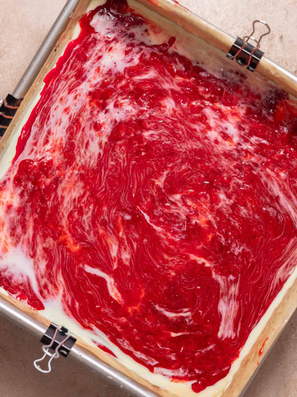 Strawberry jam swirled over cheesecake filling in a square pan.