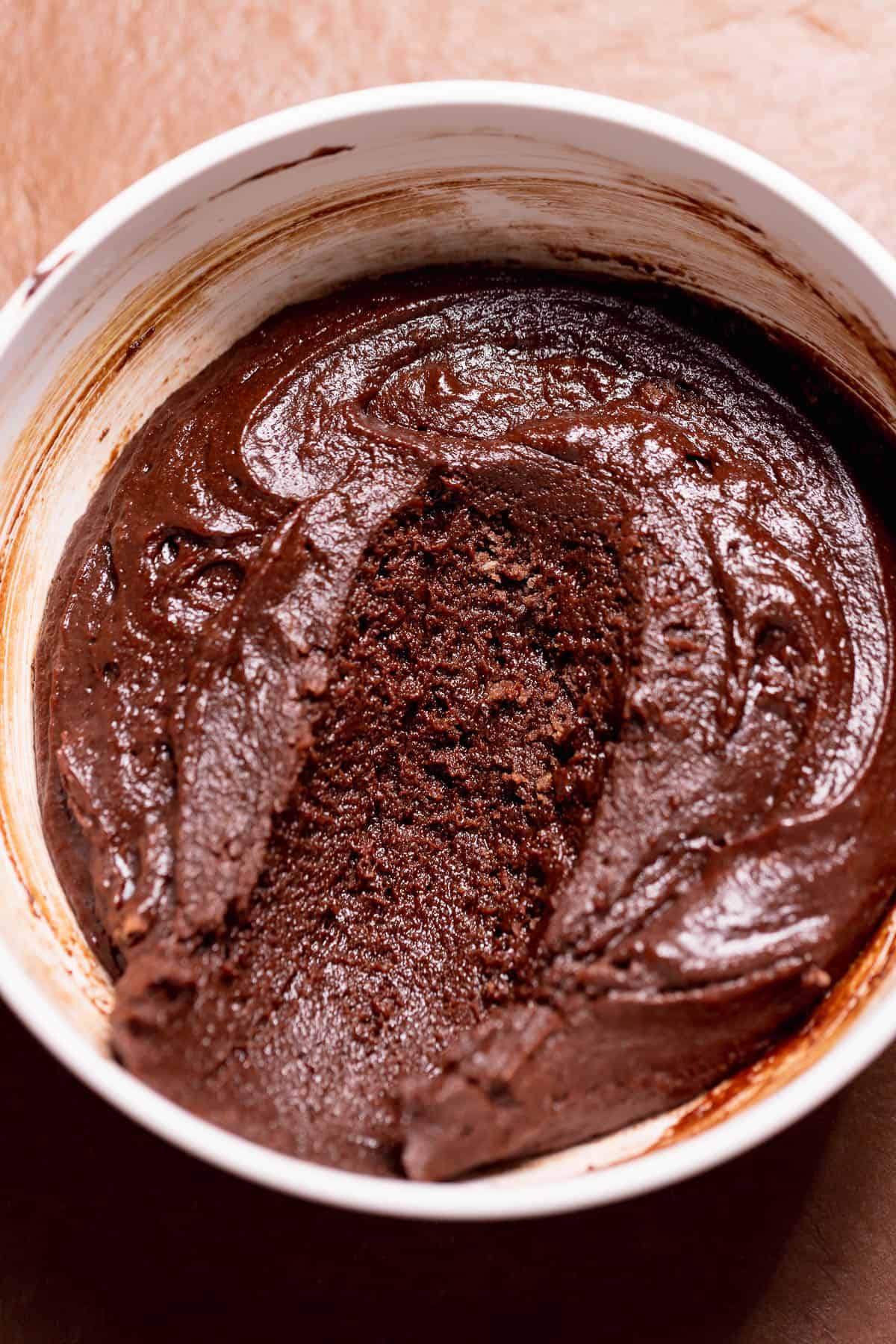 Homemade brownie batter filling in a white bowl with a spoon scoop removed, showing its thick texture.