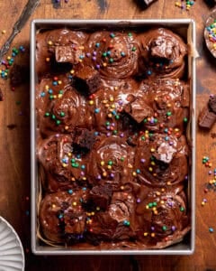 Cosmic brownie rolls topped with chocolate cream cheese frosting, fudgy brownie chunks, and rainbow candy-coated chips in a square baking pan.