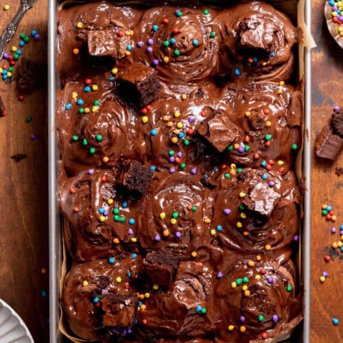 Cosmic brownie rolls topped with chocolate cream cheese frosting, fudgy brownie chunks, and rainbow candy-coated chips in a square baking pan.