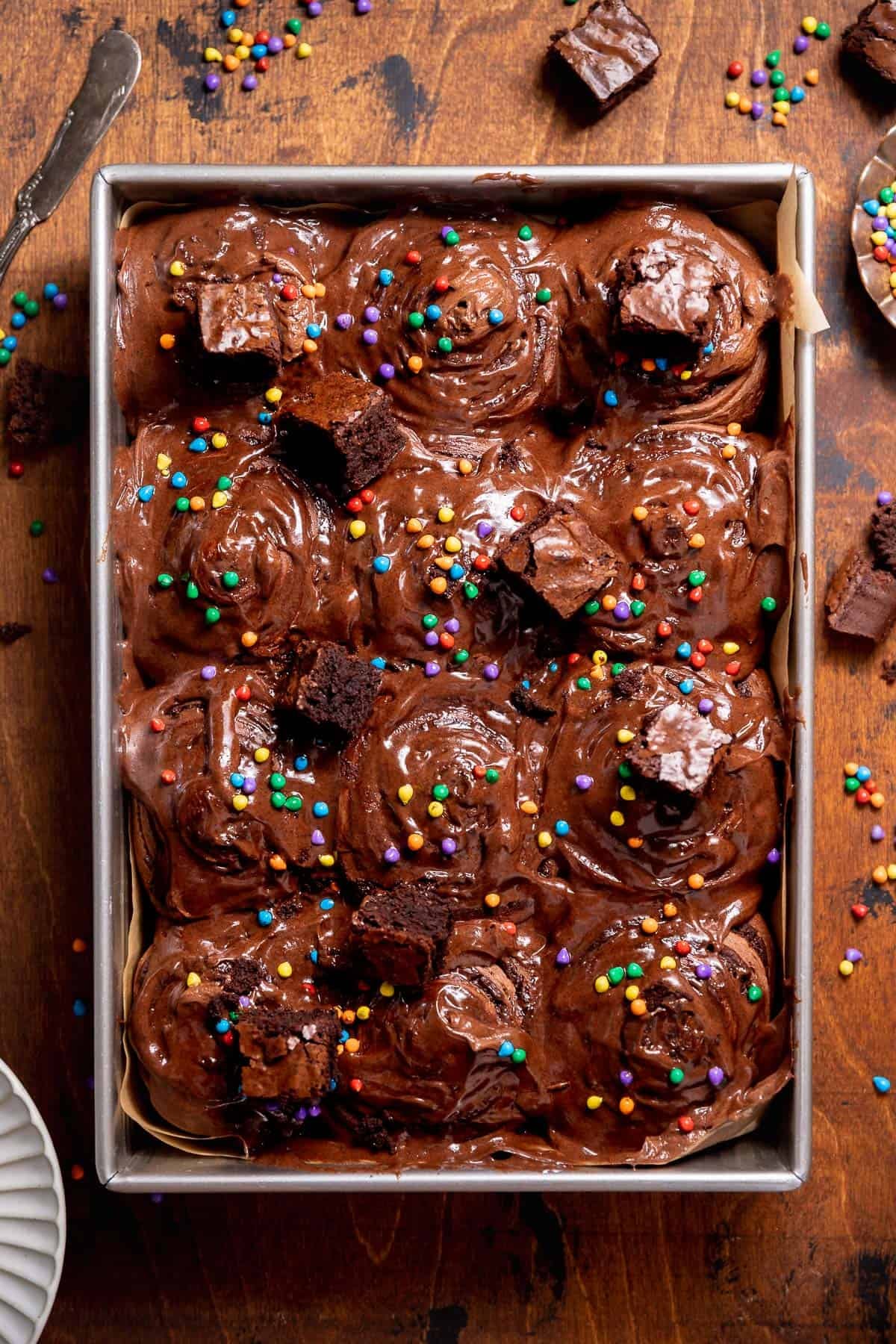 Overhead view of frosted cosmic brownie cinnamon rolls topped with brownie pieces and rainbow candy-coated chips.