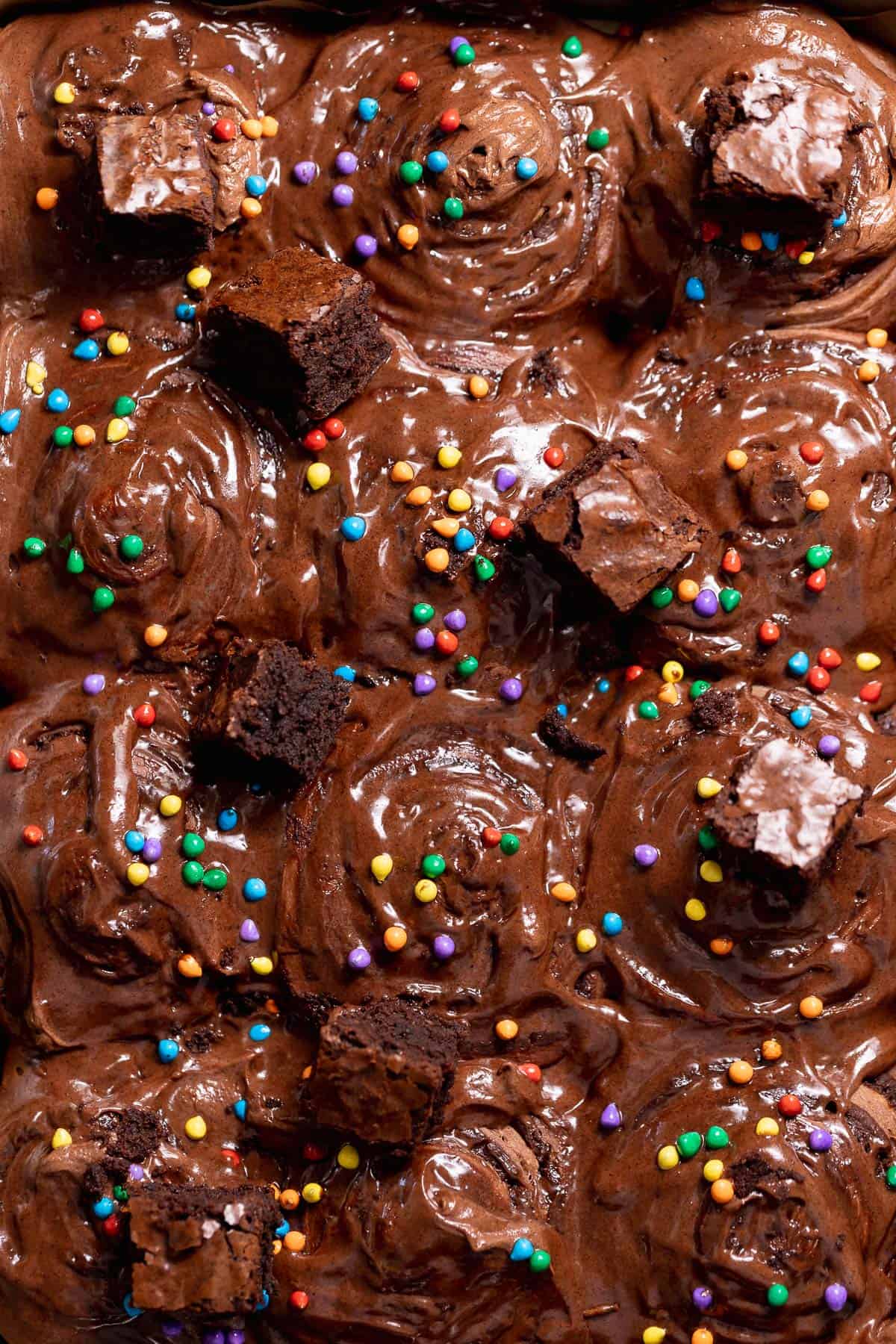 Cosmic brownie rolls topped with chocolate cream cheese frosting, fudgy brownie chunks, and rainbow candy-coated chips in a baking pan.