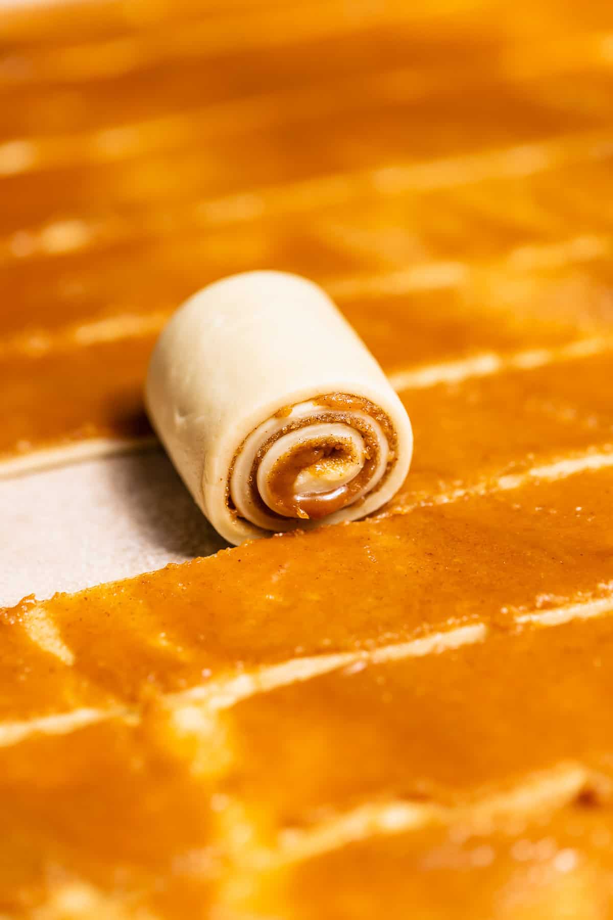 Cinnamon roll dough filled with caramel and cinnamon sugar cut into strips and being rolled up.