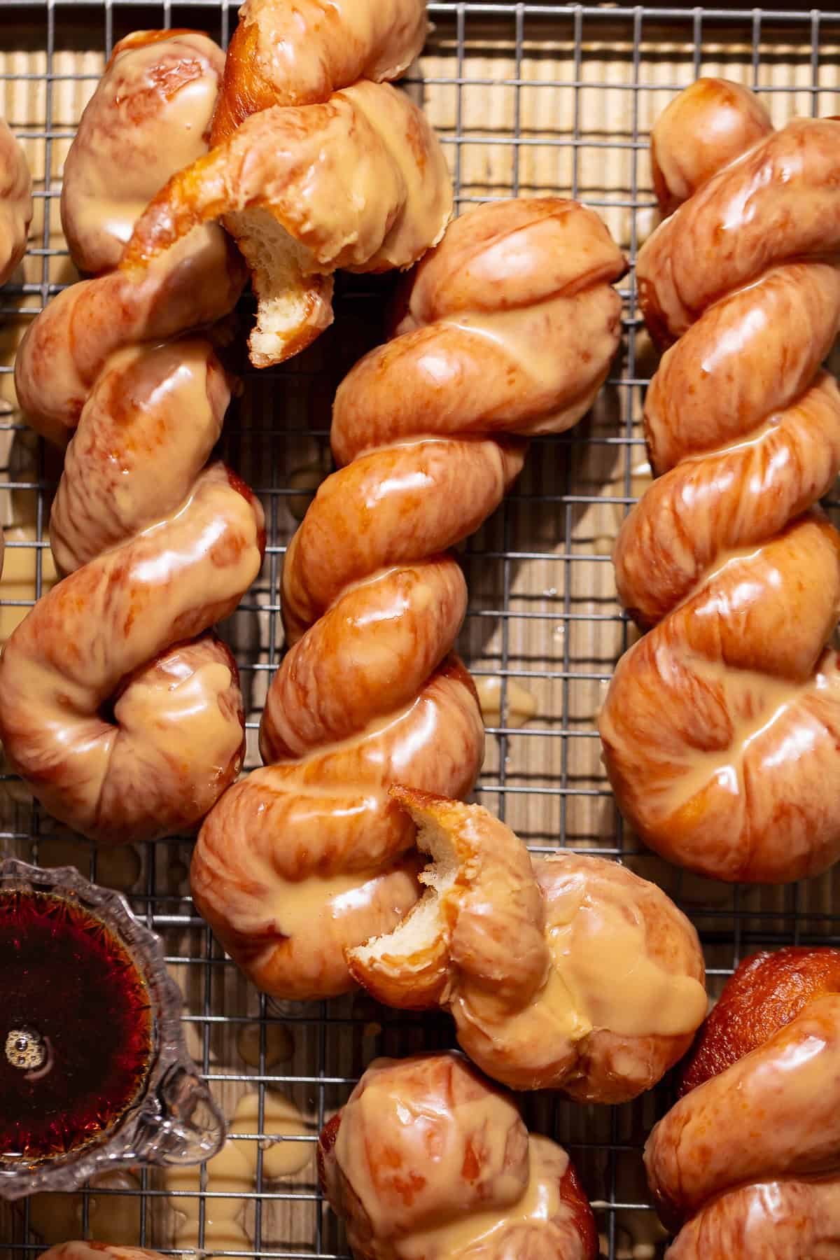 Maple glazed donut twists arranged on a wire cooling rack with a small dish of maple syrup.