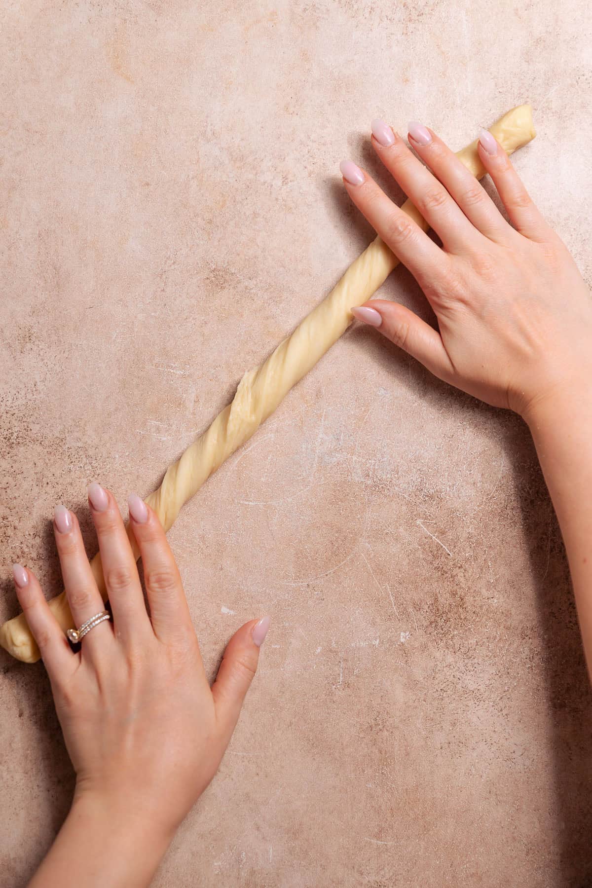 Hands rolling and twisting a long strip of donut dough on a work surface.