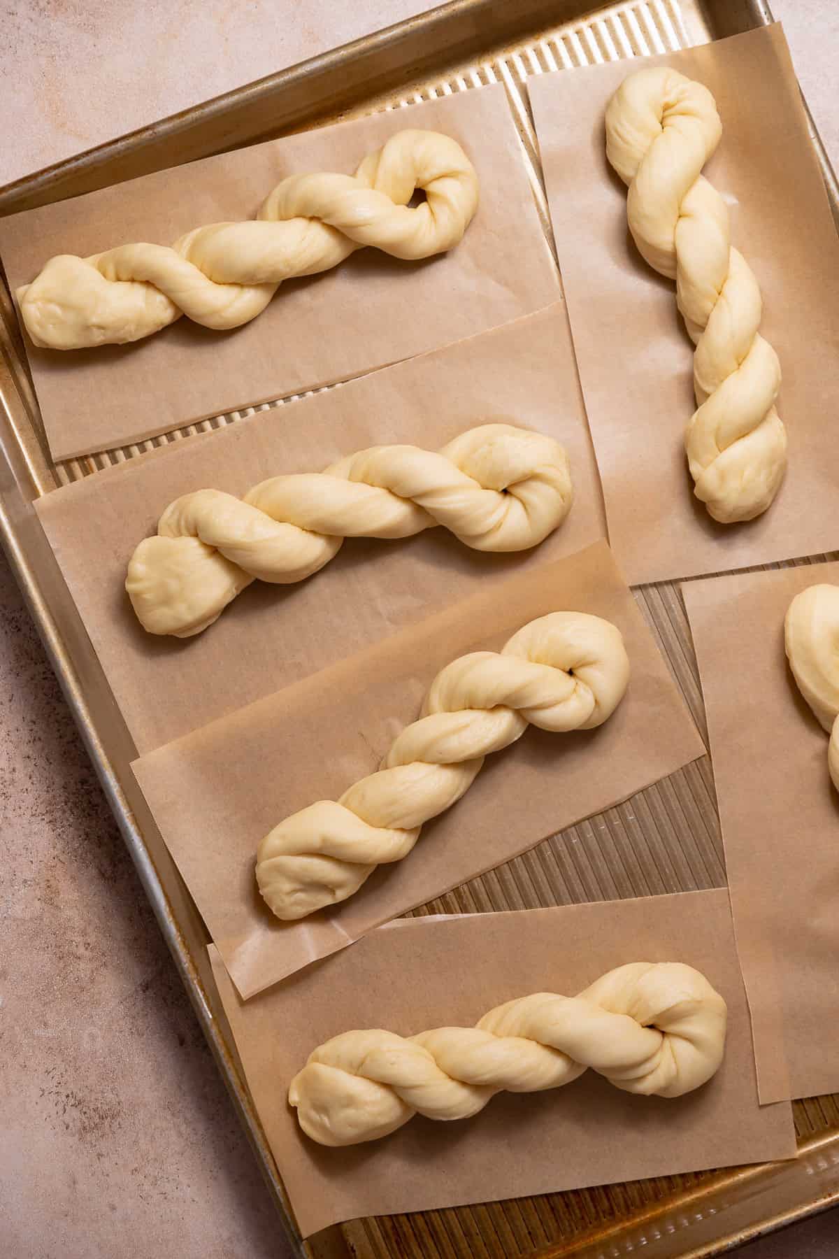 Shaped donut twists proofing on individual parchment squares on a sheet pan.