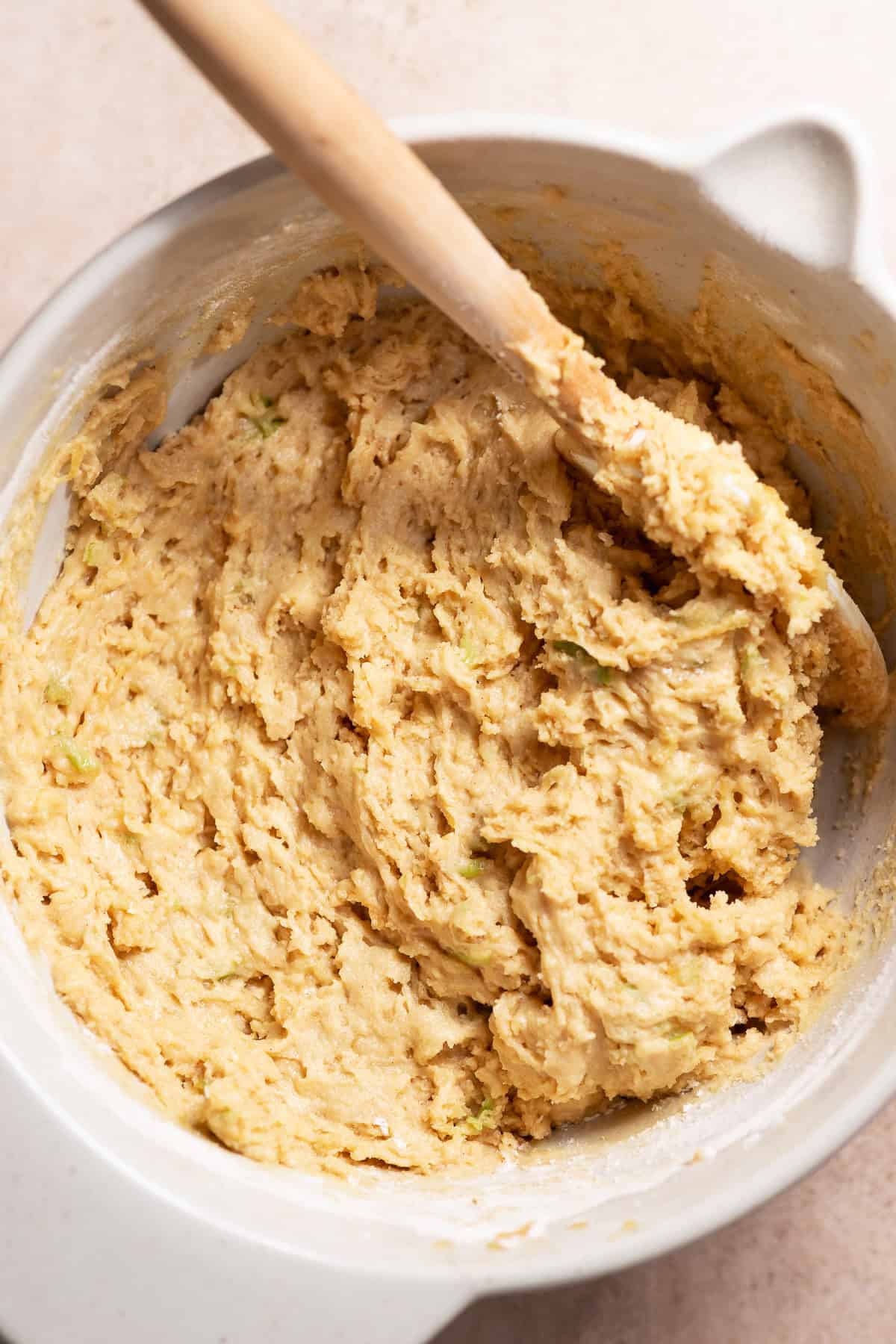 Thick apple cider donut batter with shredded apple being mixed in a bowl with a wooden spoon.