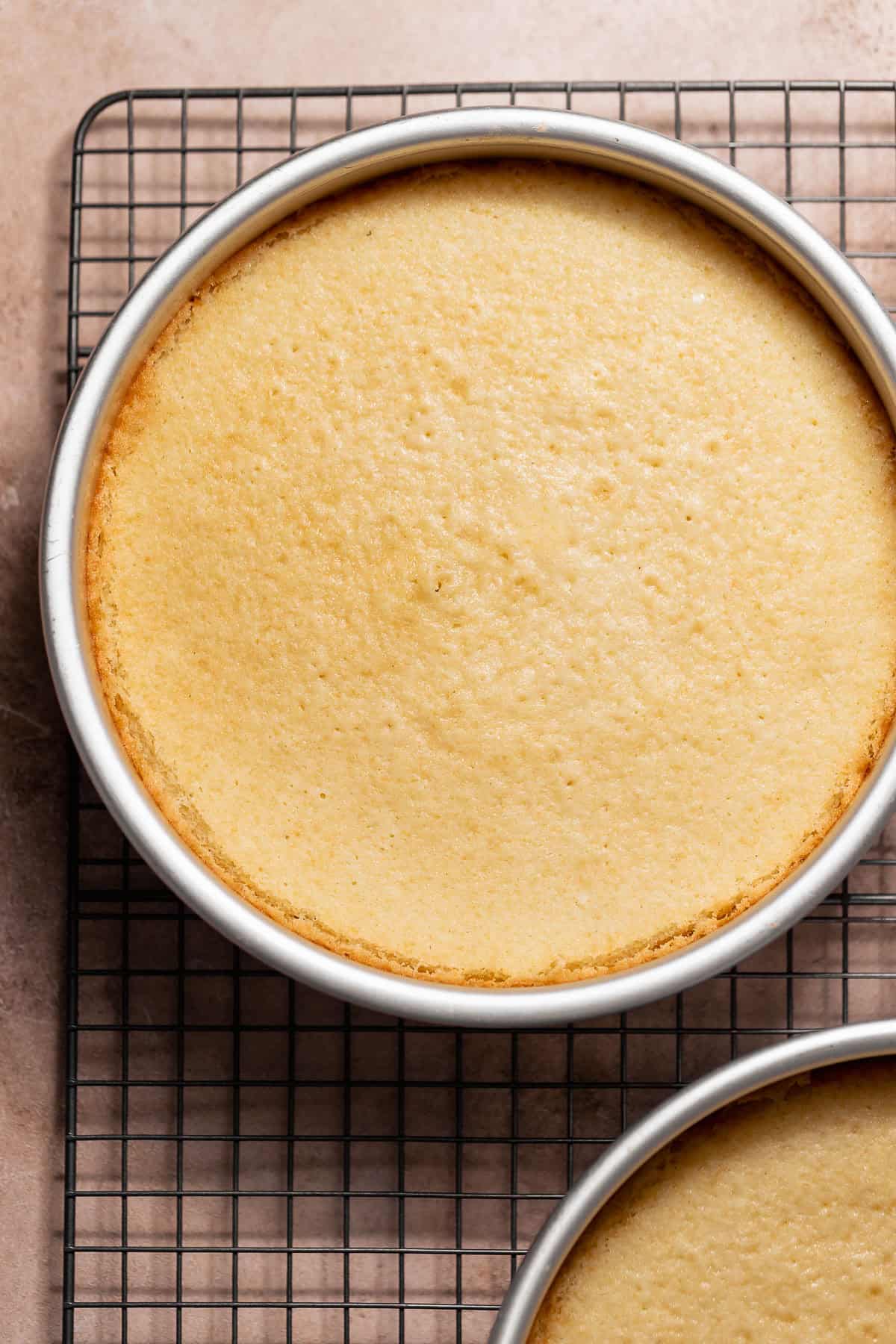 Overhead view of a golden vanilla cake layer in a round metal pan on a wire cooling rack.