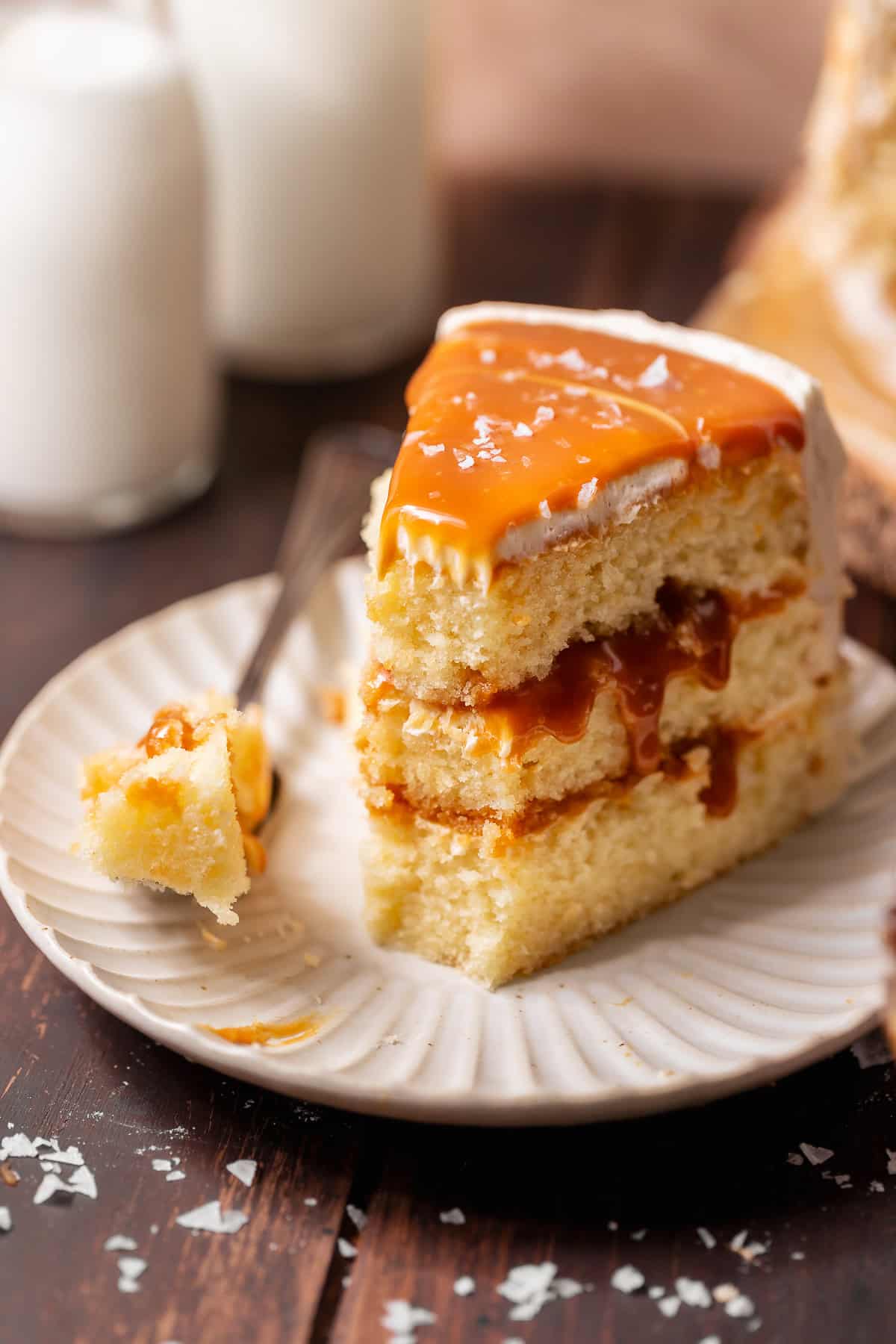 Slice of three-layer caramel cake on a white plate with a fork holding a bite of cake, caramel filling, and frosting.