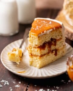 Slice of caramel cake on a white plate with three vanilla cake layers, caramel filling, buttercream frosting, and a fork beside it.