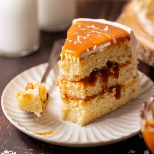 Slice of caramel cake on a white plate with three vanilla cake layers, caramel filling, buttercream frosting, and a fork beside it.