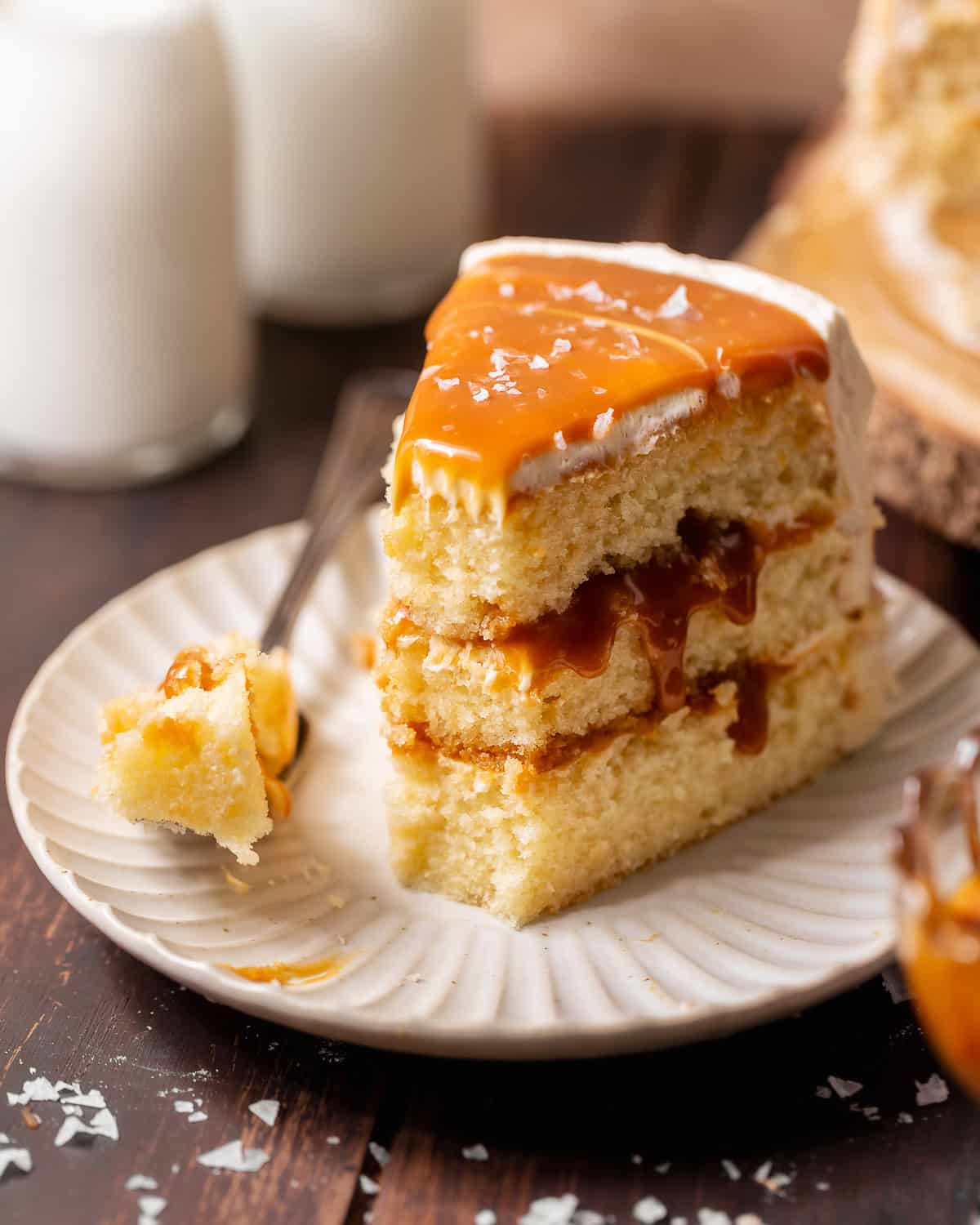 Slice of caramel cake on a white plate with three vanilla cake layers, caramel filling, buttercream frosting, and a fork beside it.