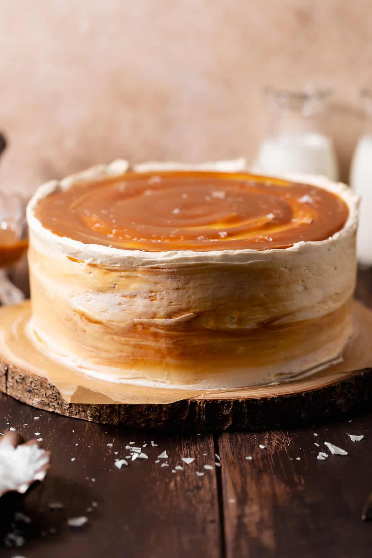 Whole caramel layer cake with swirled caramel buttercream frosting, glossy caramel topping, and flaky sea salt on a wooden cake stand.