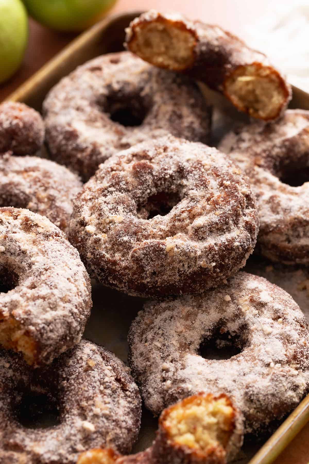 Cinnamon sugar apple cider donuts piled on a baking sheet with broken pieces showing the soft interior.