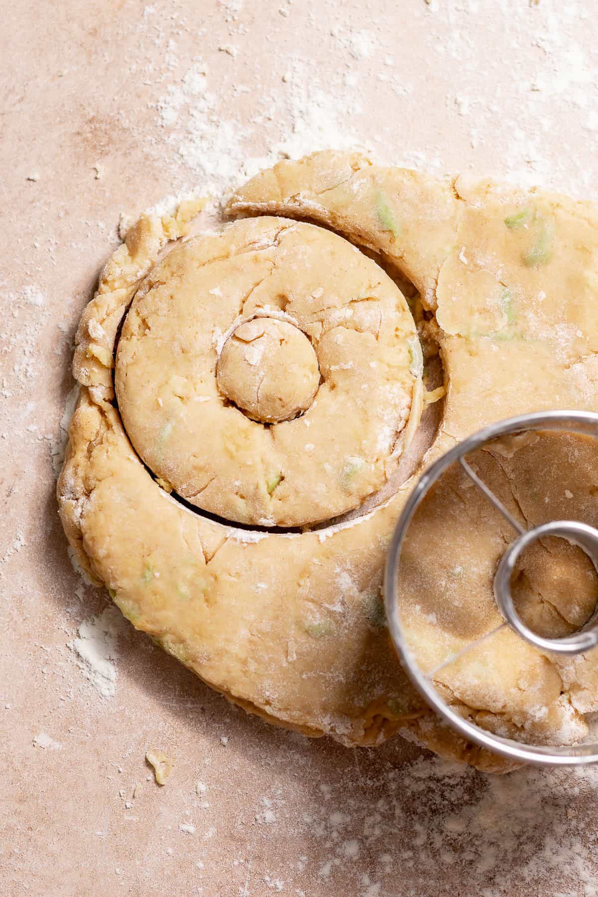Rolled apple cider donut dough being cut into rings with a metal donut cutter on a floured surface.