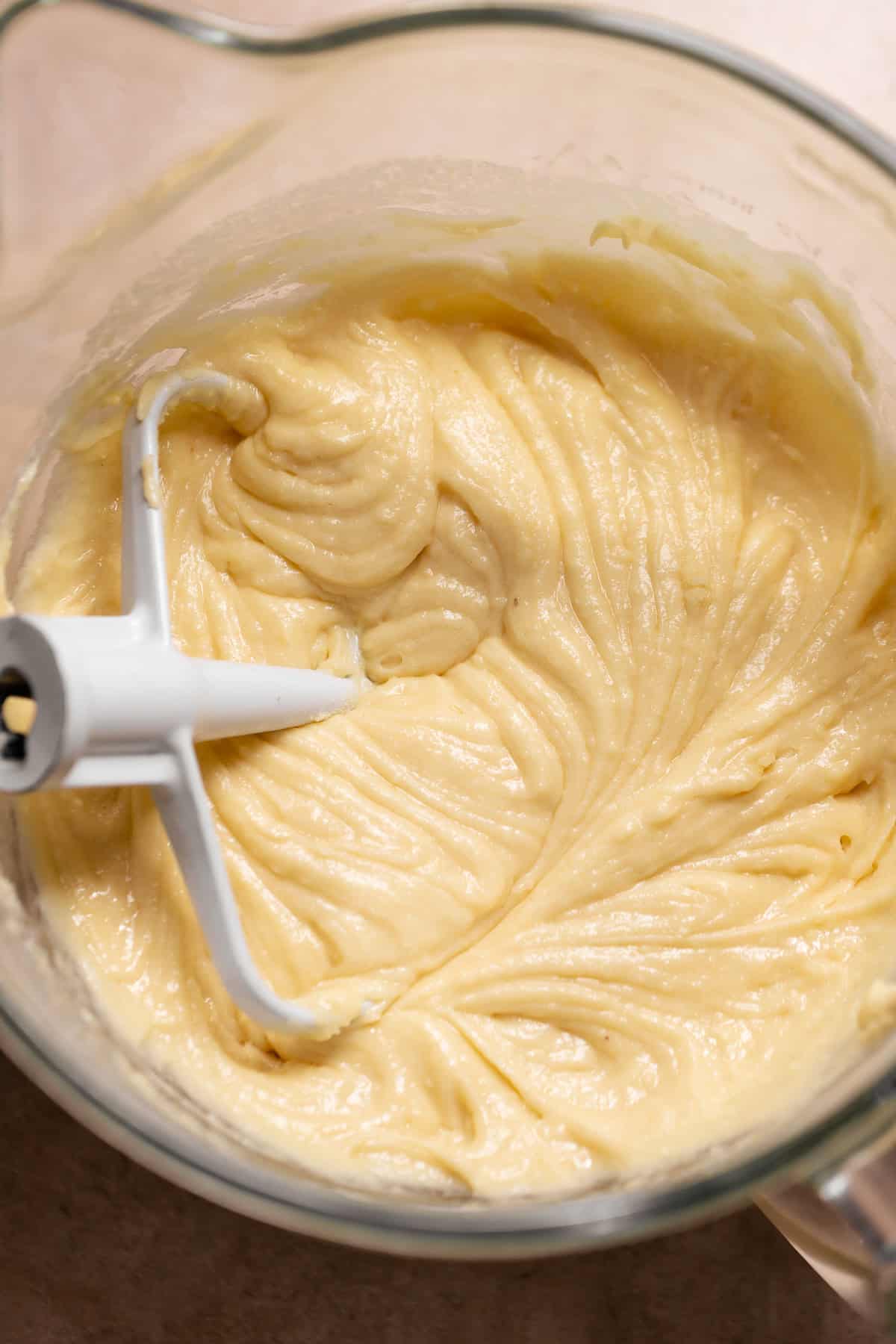 Close-up of thick, pale yellow vanilla cake batter being mixed with a white paddle attachment in a glass mixing bowl.
