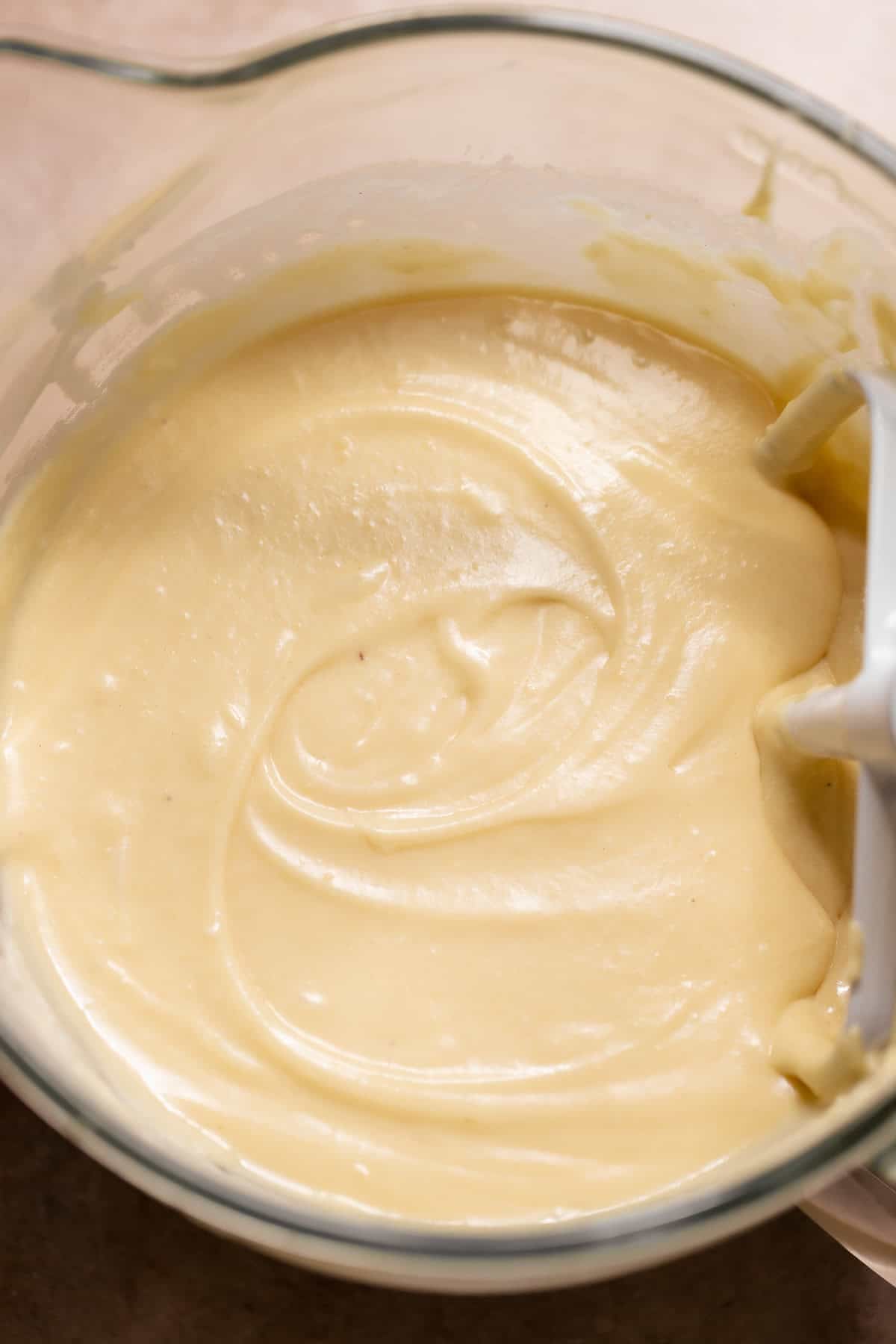 Close-up of smooth, pale yellow vanilla cake batter in a glass mixing bowl with a white paddle attachment resting along the side.