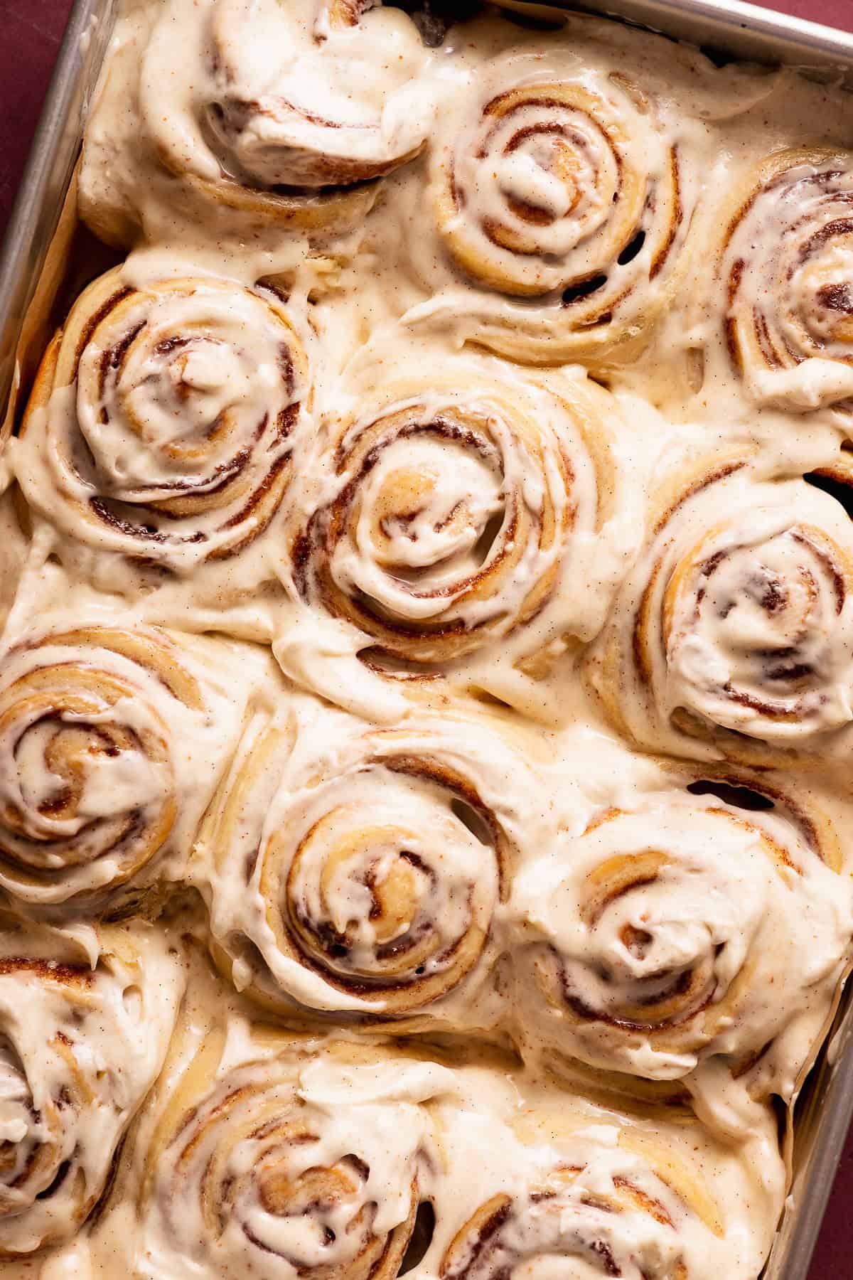 A pan of freshly baked cinnamon rolls topped with a thick layer of white cream cheese icing speckled with brown butter solids.