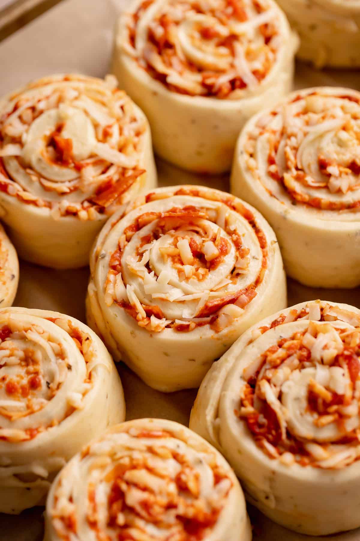 Soft, risen pepperoni pizza rolls with visible spirals of cheese and tomato sauce, arranged closely together before baking.
