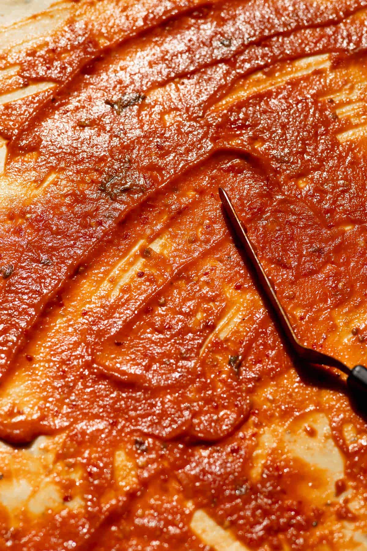 A metal offset spatula spreading red pizza sauce over a layer of dough.